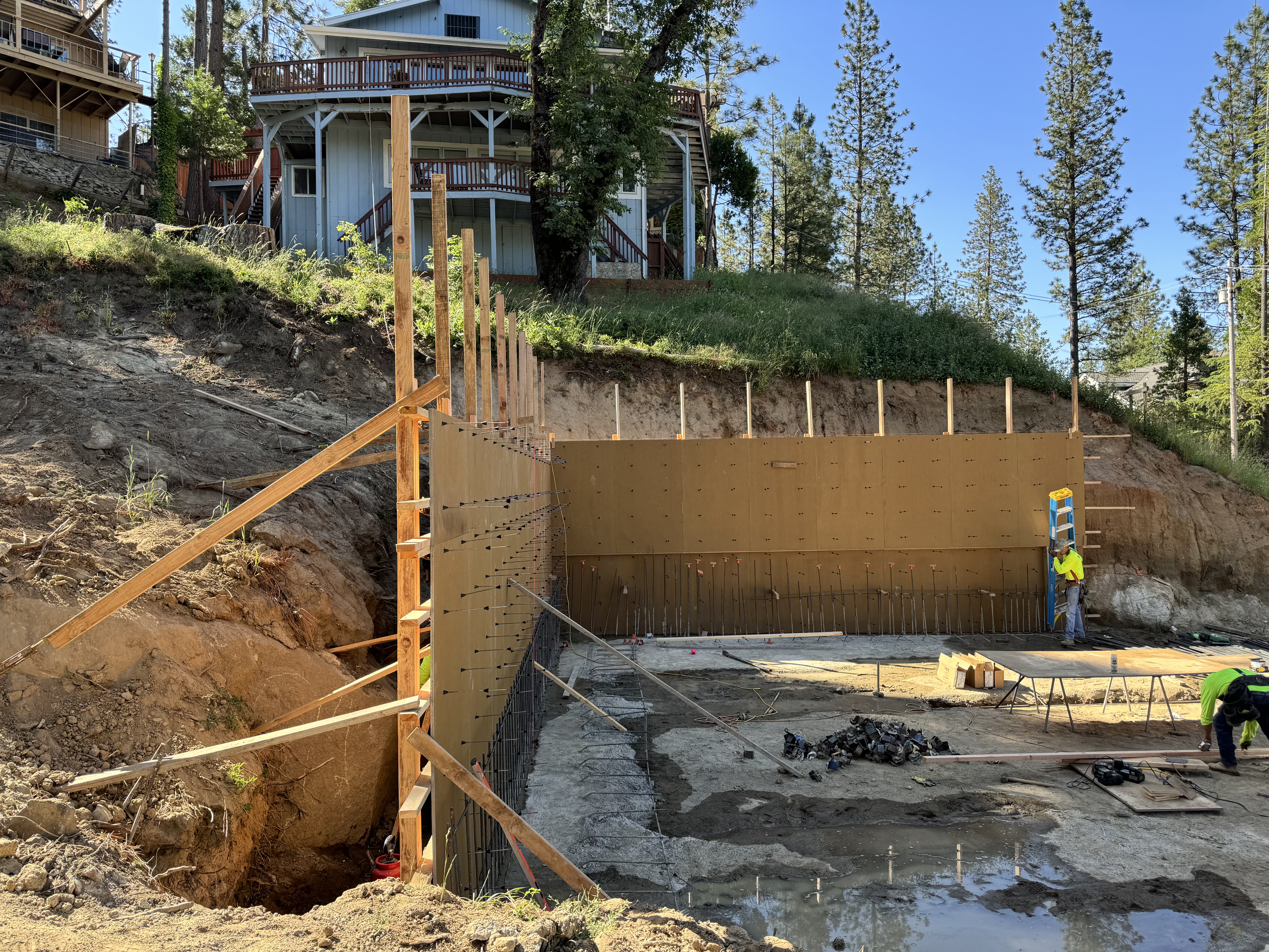 Large concrete retaining wall formwork construction on steep hillside with house above